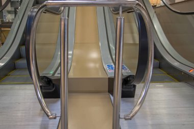 Asansörün önünde krom çitli, şeffaf cam kenarlı ve merdivenlerin önünde metal tırtıklı platformları olan, bej merkezi bir kısmı olan yürüyen merdivenin alt kısmı..