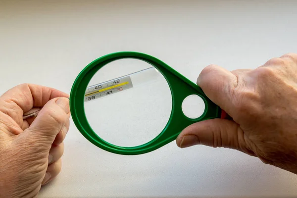 Elderly woman holding with left hand thermometer that shows 40 degrees Celsius, in right hand green magnifying glass magnifies measurement. Concept: High temperature, harbinger of flu or coronavirus