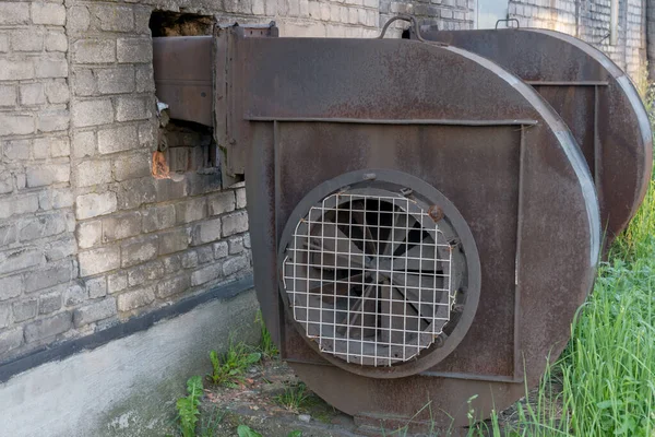 Industrial centrifugal radial exhaust fans of snail type of brown color with traces of rust, installed near building made of white silicate bricks with traces of soot, at side of which grass grows