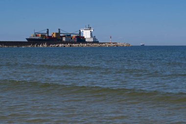 Mavi gövdeli kargo gemisi çeşitli renklerde nakliye konteynırlarıyla dolu. Kıyıdaki körfeze giriyor. Kenarında bir deniz feneri ve yanında açık mavi bir gökyüzüne karşı beton dalgalanması olan bir rıhtımdan geçiyor.