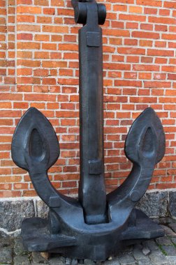 Gri bir taşın üzerinde duran iki büyük sivri bacaklı, dev bir antika siyah metal çapa, kırmızı tuğladan bir duvara tutturulmuş ve desteklenmiş.