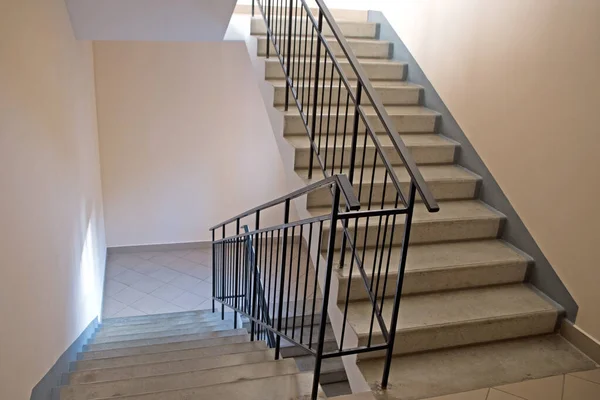 Bir apartmanın girişindeki merdivenler. Beton basamaklı iki kat merdiven ve bej duvarlar boyunca siyah metal korkuluklar, alttaki inişler büyük gri fayanslarla kaplı.
