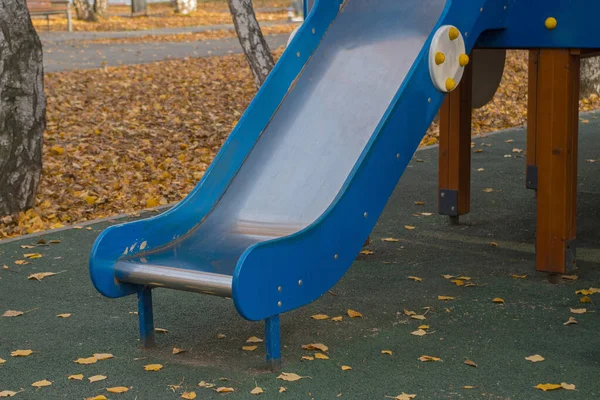 金属コーティングを施した木製のスライド 塗装された青 白樺の木と秋の日に黄金の落ち葉で公園内の遊び場の複合体内の緑の滑り止めコーティングに立っています — ストック写真