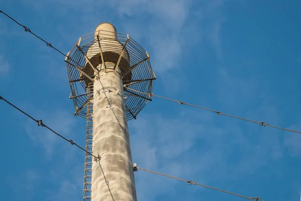 View from below of asbestos-cement boiler house pipe installed vertically, supported by system of metal tripwires, with observation platform at top and ladder of brackets rising up against cloudy sky