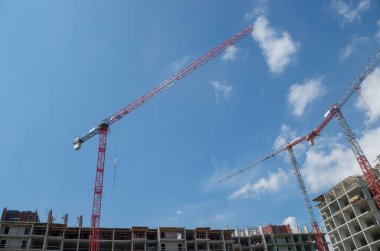 İnşaat alanındaki kırmızı kule vinçleri, inşaat halindeki apartmanların gri çerçevelerinin üst katlarına oklarını seriyorlar..