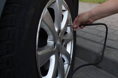 Gri asfalt bir otoparkın kaldırımında çömelmiş bir kadın açık mavi bir arabanın ön siyah tekerleğini siyah bir kompresörle pompalıyor. Kavram: kadın pis adamın işini yapıyor.