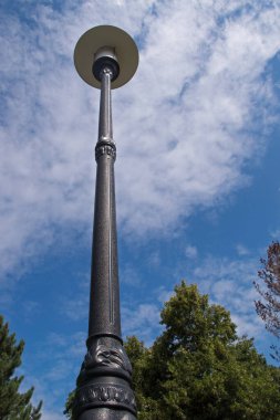Bir sokak lambasının yuvarlak beyaz abajuru yüksek bir direkte siyah metalle süslenmiş mavi gökyüzüne karşı levrek ve yeşil ağaçlarla çevrili lekeli kıvırcık bulutlar.