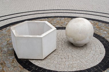 Beyaz beton üç boyutlu geometrik şekiller içi boş bir altıgen ve siyah bir kaldırım dairesi içinde gri, bej ve siyah fayanslarla kaplı bir platformda duran katı bir top.