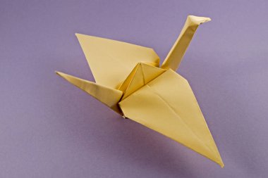 Origami, sarı kalın kağıttan yapılmış bir vinç figürü şeklinde, yukarı ve sağa doğru mor bir zemin üzerinde katlanmış. Kavram: Barış, mutluluk ve neşe için çabalama.