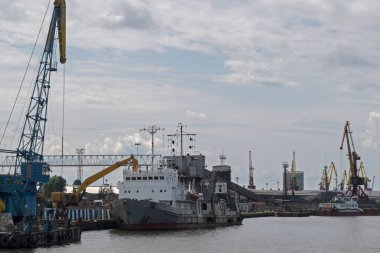 Limanda kazıcı kullanan beyaz güverteli gri bir yük gemisinin kömür yüklemesi veya boşaltılması, yakınlardaki yükleme platformları ve port yükleme vinçleri arka planda bulutlu mavi bir gökyüzü görülebilir