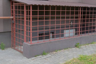 Kapısı siyah asma kilitli bir kafes, kırmızı metalden kaynaklanmış, bodruma beyaz kapı ve gaz borusuyla çevrili, kahverengi cepheli bir apartman dairesinin önünden geçiyor.