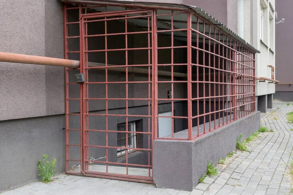 Cage with locked gate with black padlock, welded from red metal, enclosing entrance to basement with white door and gas pipe passing over it in residential apartment building with a brown facade