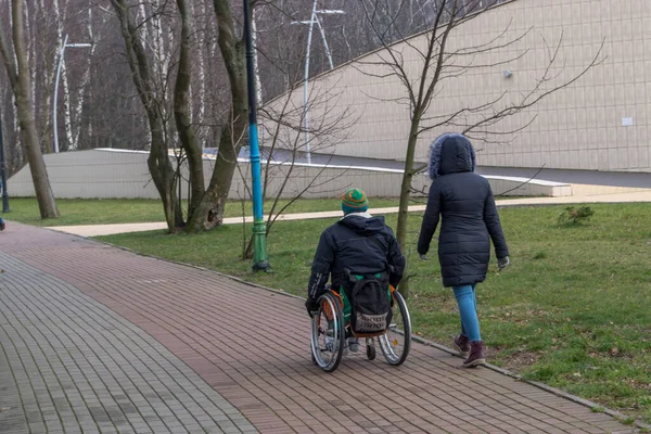 Siyah ceketli tekerlekli sandalye kullanıcısı ve sırtında sırt çantası olan yeşil şapkalı siyah ceketli kız arkadaşı ve ağaçların arasında yürümekte olan kot pantolonlu. SVETLOGORSK, RUSYA, 15 Şubat, 2020