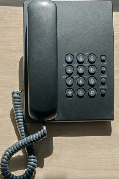 A black plastic landline phone sits on a wooden beige office Desk, illuminated by the light falling from the window, top view. Concept: business related to phone calls, helpdesk, service desk.