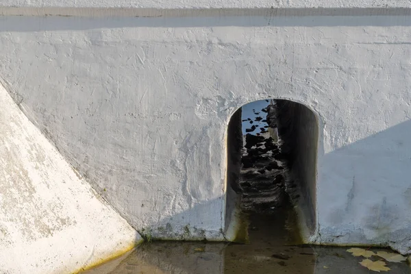 Whitewashed or painted white concrete drainage structure with hole in the form of an arch on the ground with rainwater at the bottom under road or rail surface. Concept: protecting road from drowning