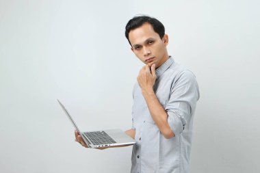 Asyalı endonezyalı genç adam elinde dizüstü bilgisayarla ciddi bir yüz ifadesiyle bir soru soruyor. Tecrit edilmiş arka planda kafa karıştırıcı bir fikir hakkında düşünceli.