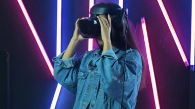 Genç bir kadın kulaklığını takıyor, etrafına bakıyor ve sanal gerçeklikte ne kadar harika olduğunu merak ediyor. Renkli ışıklandırma arka planında sanal gerçeklik miğferi