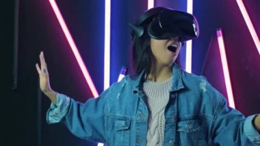 Genç bir kadın şoke olmuş, etrafına bakıyor ve VR kulaklıkla ne kadar harika olduğunu merak ediyor. Renkli ışıklandırma arka planında sanal gerçeklik miğferi