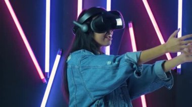 VR kulaklıkla oynayan ve gülümseyen genç bir kadın. Renkli ışıklandırma arka planında sanal gerçeklik miğferi