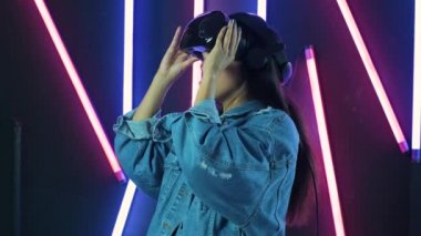 Genç bir kadın VR kulaklığı takıyor ve sanal gerçeklik dünyasında ne kadar harika olduğunu merak ediyor. Renkli ışıklandırma arka planında sanal gerçeklik miğferi