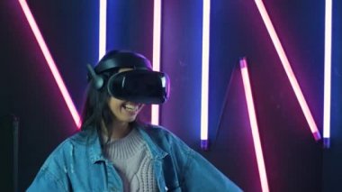 Oynayan, dans eden ve VR kulaklıkla gülümseyen mutlu genç bir kadın. Renkli ışıklandırma arka planında sanal gerçeklik miğferi