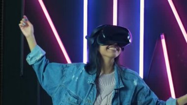 Mutlu genç kadın etrafına bakıyor ve VR kulaklıkla ne kadar harika olduğunu merak ediyor. Renkli ışıklandırma arka planında sanal gerçeklik miğferi