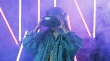 Genç bir kadın VR kulaklığı takıyor ve sanal gerçeklik dünyasında ne kadar harika olduğunu merak ediyor. Renkli duman arka planında sanal gerçeklik miğferi