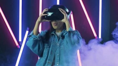 Genç bir kadın VR kulaklığı takıyor ve sanal gerçeklik dünyasında ne kadar harika olduğunu merak ediyor. Renkli duman arka planında sanal gerçeklik miğferi
