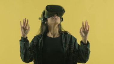 Genç bir kadın etrafına bakıyor ve VR kulaklıkla ne kadar harika olduğunu merak ediyor. Sarı arka planda sanal gerçeklik miğferi