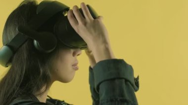 Genç bir kadın vr kulaklık takıyor, etrafına bakıyor ve sanal gerçeklik dünyasında ne kadar harika olduğunu merak ediyor. Sarı arka planda sanal gerçeklik miğferi