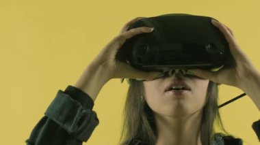 Genç bir kadın vr kulaklık takıyor, etrafına bakıyor ve sanal gerçeklik dünyasında ne kadar harika olduğunu merak ediyor. Sarı arka planda sanal gerçeklik miğferi