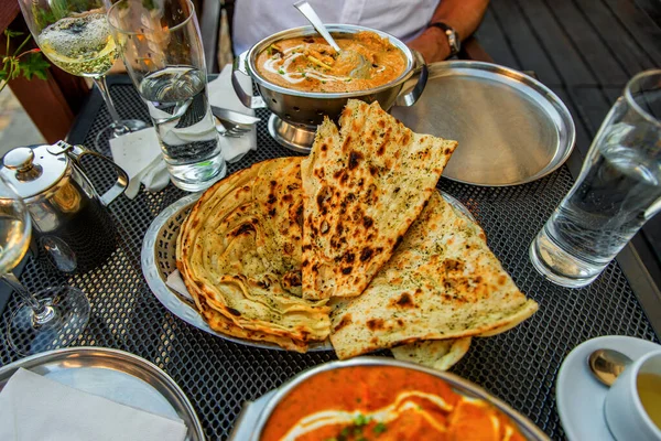Table rich of indian meals, bread (naan), glasses of water,wine,jug with tea, bowls and plate. Outdoor indian restaurant.