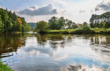 Açık nehir yüzeyinde yansıyan bulutlar, kıyılarda açık yeşil ağaçlar, sonbaharın başlangıcında güneşli bir gün. Luznice Nehri, Çek Cumhuriyeti.