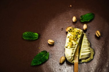 Dilimlenmiş şam fıstıklı dondurma (beyaz çikolata, ezilmiş fındık ve şam fıstığı dolgusu), fındık ve nane yapraklarıyla aydınlatılmış dairede, çiğ kahverengi arka planda eriyor..