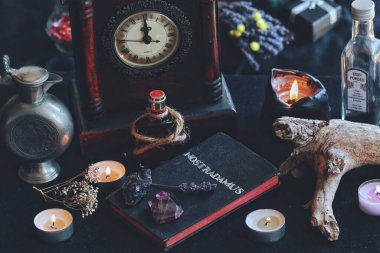 Hand made book with silver foil word Nostradamus written on it, about predicting the future and ancient Nostradamus prophecies. Witchy vibe background, filled with burning lit candles, dried herbs, flowers, jars, bottles, old clock tree bark clipart