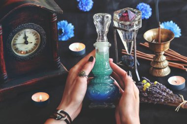 Wiccan cadısı, mihrabında bir iyi geceler iksiri tutuyor. Lavanta, tütsü yakıcı, eski saat ve kristallerle bulanık arka planı olan güzel bir şarap şişesinde mavi renkli sıvı.