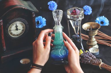 Wiccan cadısı, mihrabında bir iyi geceler iksiri tutuyor. Lavanta, tütsü yakıcı, eski saat ve kristallerle bulanık arka planı olan güzel bir şarap şişesinde mavi renkli sıvı.