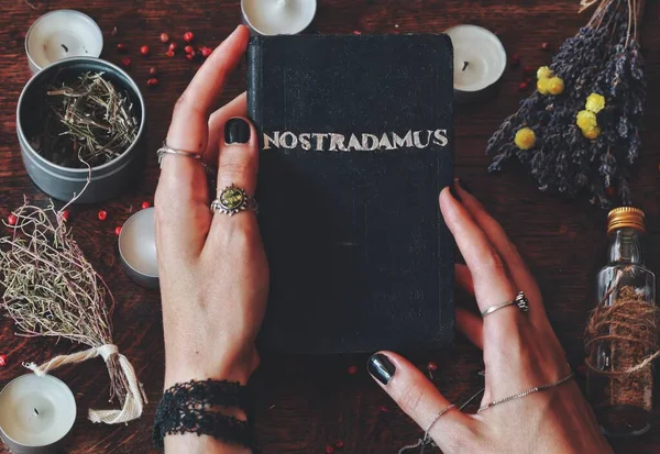 Wiccan witch holding hand made book with silver foil word Nostradamus written on it, about predicting the future and ancient prophecies in her hands. Witchy background filled with unlit candles, dried herbs flowers, jars bottles, old clock, tree bark