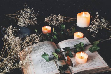 Open vintage poetry book on a black table surface with white lit burning candles and dried flowers. Dark, romantic and cozy feel with a branch of ivy on top of the pages. With light overlay of white speckles clipart