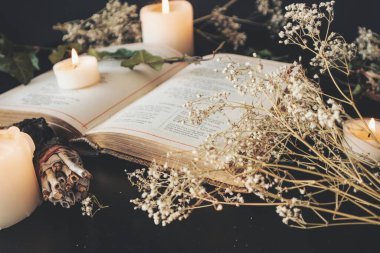 Close up of open vintage poetry book decorated with dried baby's breath flowers. Blurred background with white lit burning candles, plants and small sage stick. Black table surface. Romantic soft feel clipart