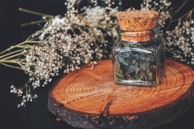 Kare cam bir kavanozdaki kurutulmuş ateş yosunu (willowhere erb) bitkilerini kapatın. Ağaç gövdesi platformunda şişe, kurumuş bebek nefesi çiçekleri siyah, biraz bulanık arka planda