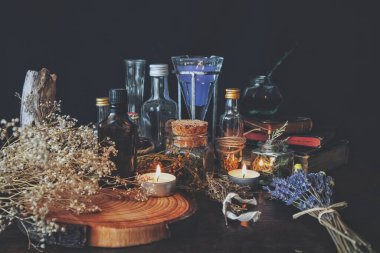 Wiccan cadı eczacısı. Sihir için çeşitli malzemeler, iksirler, kurutulmuş bitki şişeleri ve kavanozlar sunağa konur. Siyah arka plan, bebek nefesli çiçekler, lavanta, yanan beyaz mumlar, eski kitaplar ve ağaç gövdesi dilimleri.
