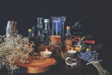 Wiccan witch apothecary - various ingredients, potions and dried herb bottles and jars for magick, placed on an altar. Dark, black background with baby's breath flowers, lavender, burning lit white candles, vintage books and a wooden tree trunk slice clipart