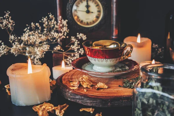 魔女のキッチンでお茶を淹れる 黒いテーブルの上の木製のプラットフォームに置かれたヴィンテージの赤白と金の色のティーカップ 背景に白いろうそくを燃やす茶 ハーブやスパイスとキッチン魔女 — ストック写真