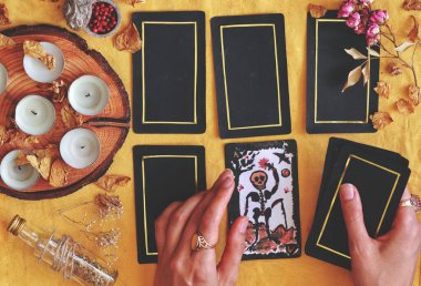Wiccan cadısı, açık sarı kumaş üzerine yayılmış 5 kart tarot kullanarak geleceği okuyor. El boyaması Ölüm tarot kartı. Kadının elinde beyaz mumlar, kurumuş çiçekler ve arka planda bitkiler var.