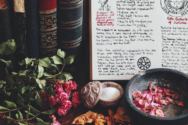 Wiccan witch altar with an open book of shadows with hand written spell in it, ready for spell casting. Grimoire page with symbols and drawings. Old vintage books, dried flowers, salt ingredients in the background
