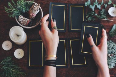 Wiccan cadısı geleceği görmek için tarot kartı okuyor. El ele tutuşan kadın eli tarot destesi. Altın kenarlı siyah kartlar. Doğa hep yemyeşil, bitkiler koyu ahşap arka planda
