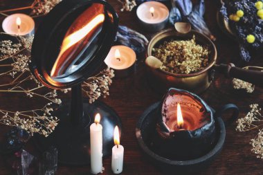Wiccan cadı sunağındaki çukur aynasında mum alevi kehaneti. Kurumuş otlar, çiçekler, kristaller, bitkiler ve karanlık arka plandaki diğer doğa nesneleriyle dolu dağınık bir masada yanan siyah mum.