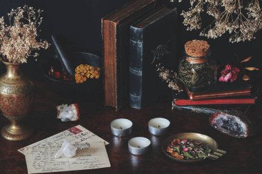 Wiccan cadı sunağındaki eski bir kitap koleksiyonu. Siyah masaya konulan eskimiş kitaplar, doğa temalı eşyalar, kurutulmuş bitkiler, çiçekler, kristaller, eski kartpostallar, havan topu beyaz mumlar gibi.