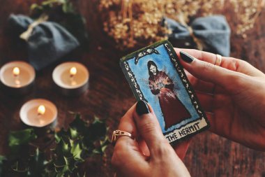 Wiccan cadısı elinde Ermiş Tarot kartı ile küçük bir el boyaması tutuyor. Kurumuş çiçekler, sarmaşık, kutsal bitkiler, yanan üç mum ve sıcak Noel gündönümü (Noel) arka planı